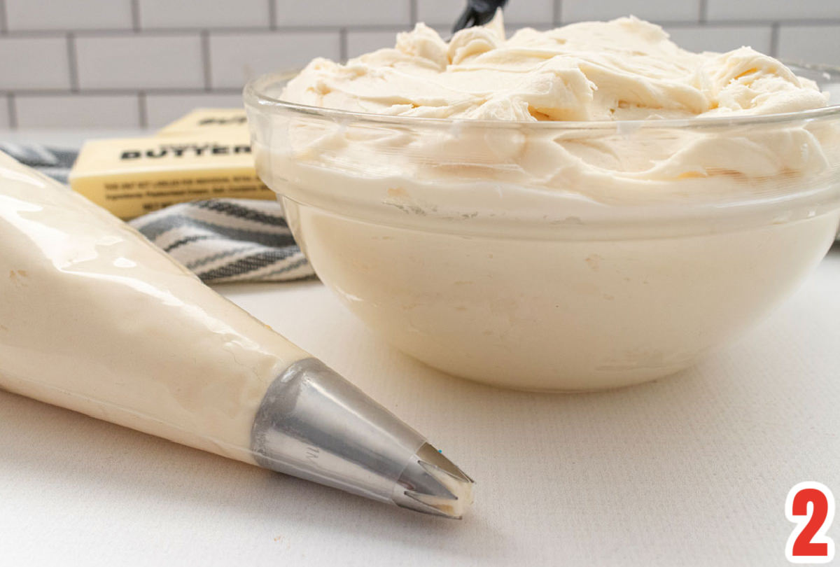 Closeup on a clear glass bowl filled with buttercream frosting sitting next to a decorating bag filled with buttercream frosting.