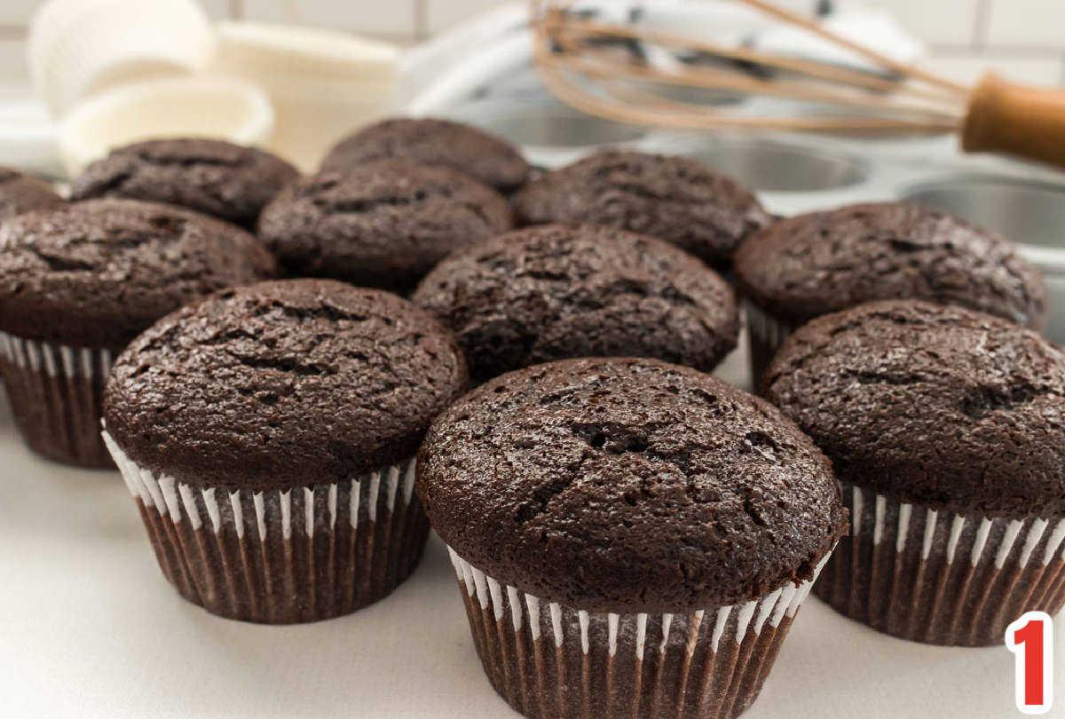 Twelve chocolate cupcakes in white cupcake liners sitting on a white surface in front of a table linen, a whisk and white cupcake liners.