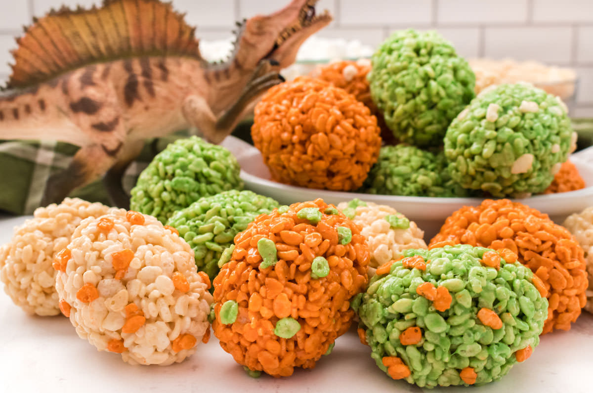 Dinosaur Eggs Rice Krispie Treats in a  white bowl and spilling out of the white bowl in front of a toy dinosaur.