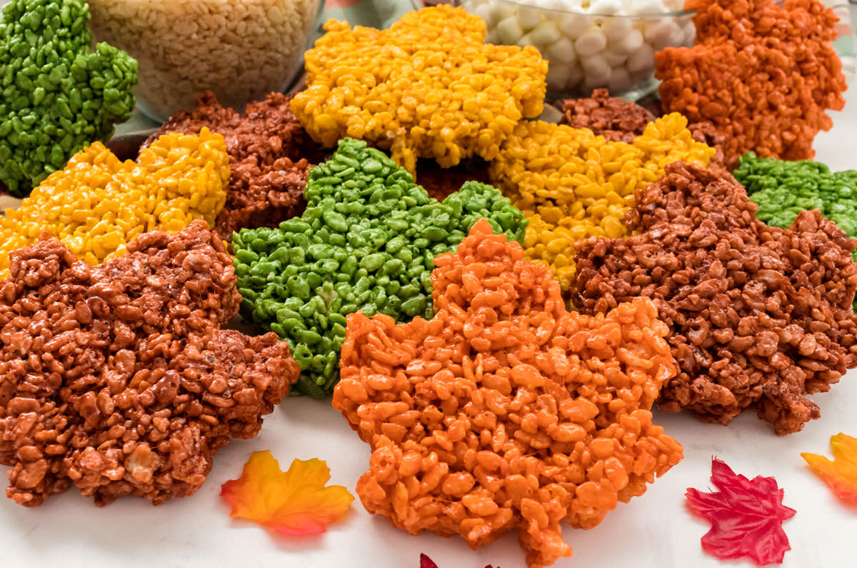 Closeup on a dozen Fall Leaves Rice Krispie Treats laying on a white table in front of glass bowls filled with mini marshmallows and Rice Krispie Cereal.