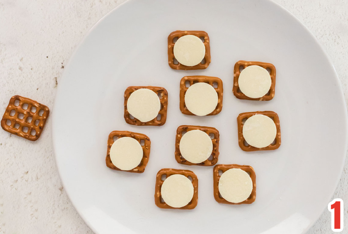 Close up on white plate sitting on a white table covered with pretzel snaps and white candy melts.