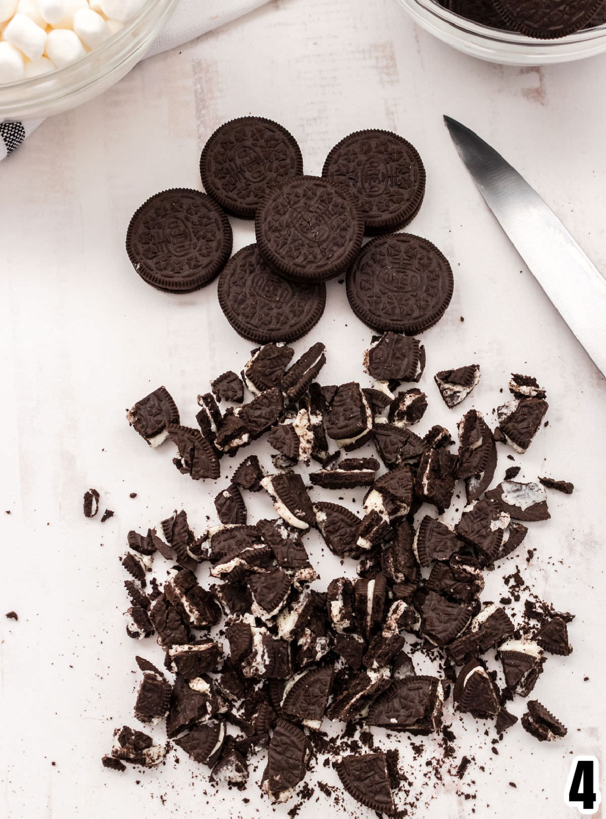 Closeup of chopped up Oreo Cookies laying on a white surface with whole Oreo Cookies and a knife.