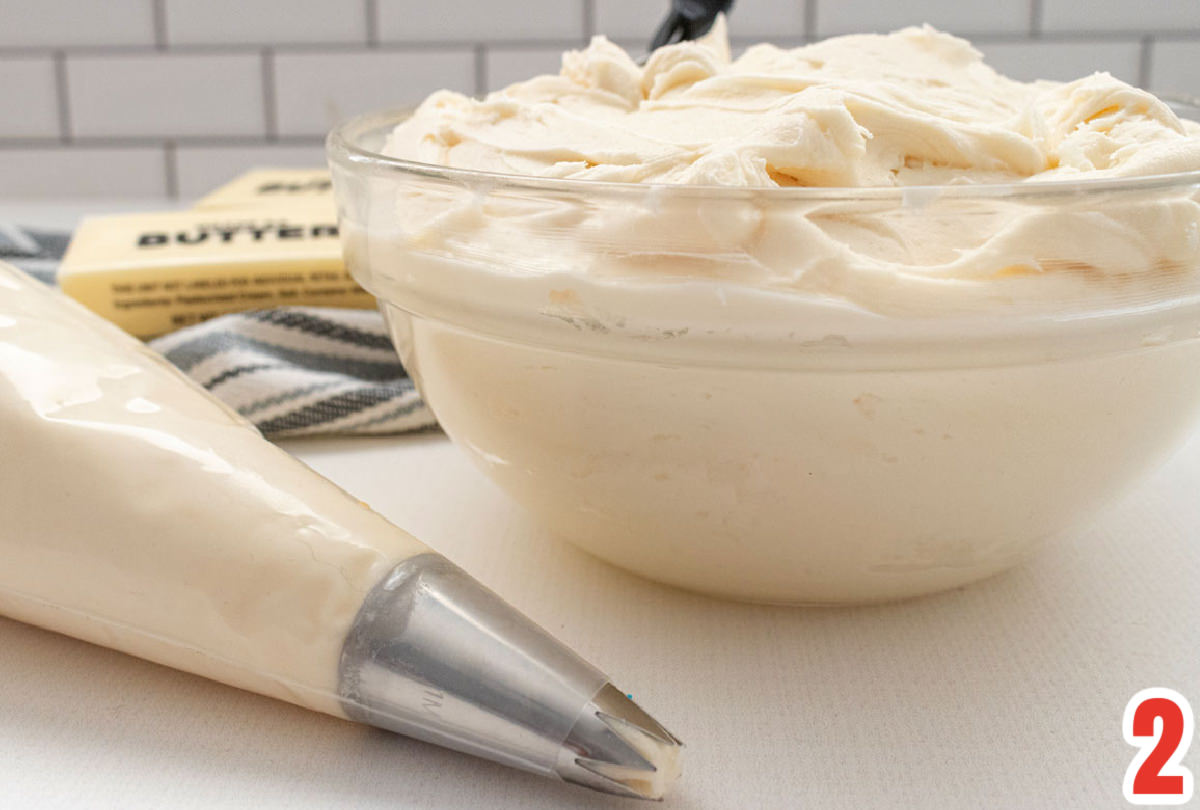 Closeup on a glass bowl filled with homemade Buttercream Frosting sitting next to a decorating bag filled with Buttercream Frosting.