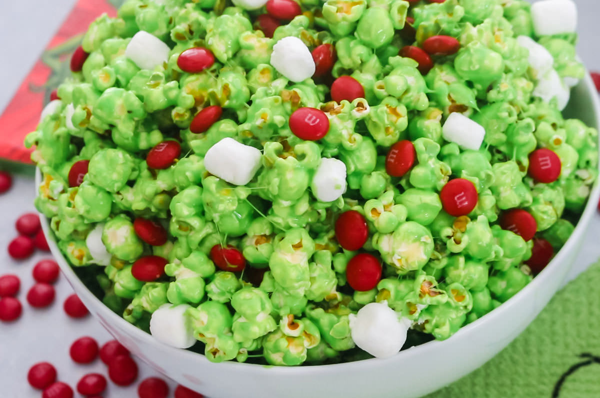 Closeup on a white serving bowl filled with Grinch Popcorn sitting on a grey table surrounded by Red M&M's and a grinch kitchen towel.