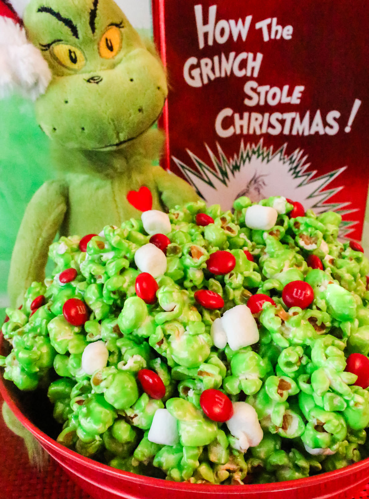 Closeup on a red bowl filled with Grinch Popcorn sitting in front of a Grinch stuffed animal and a copy of the book, How the Grinch Stole Christmas.