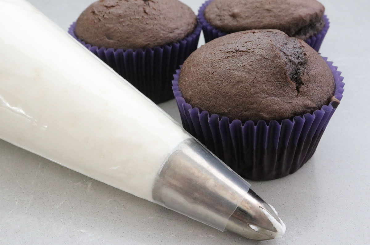 Closeup on a decorating bag filled with Whipped Cream Frosting sitting next to three unfrosted chocolate cupcakes.