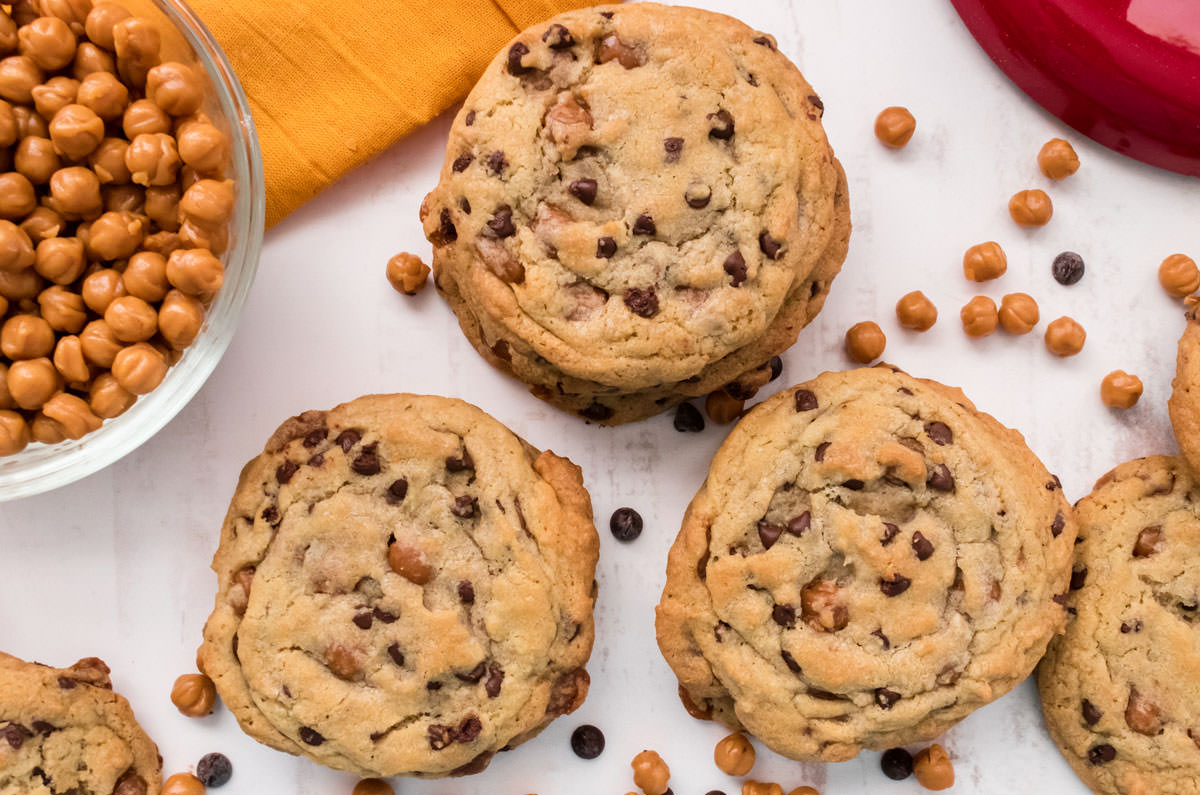 Closeup on three stacks of Caramel Chocolate Chip Cookies sitting on a white table alongside of a glass bowl filled with Caramel Bits.