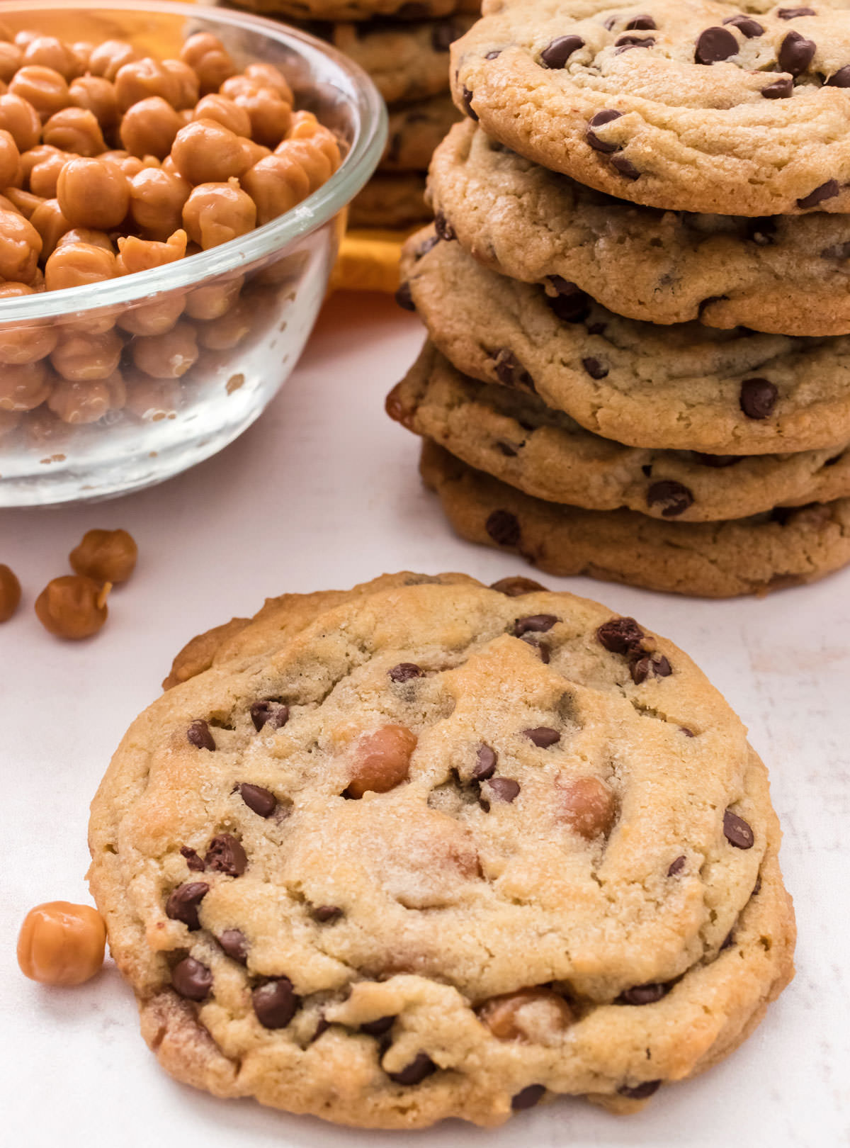 Closeup on a single Caramel Chocolate Chip Cookie sitting on a white surface in front of a stack of cookie and a glass bowl filled with Kraft Caramel Bits.