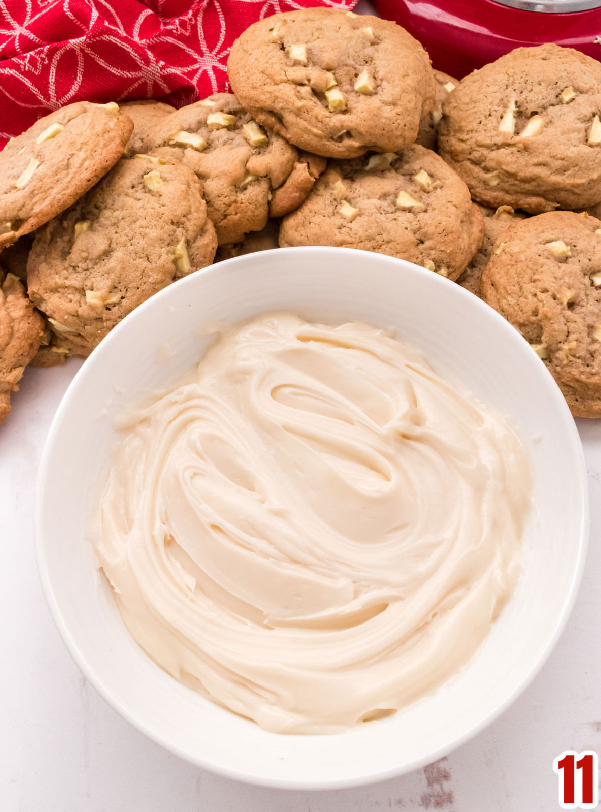 Closeup on a white bowl filled with Homemade Cream Cheese Frosting surrounded by the Apple Cookies.