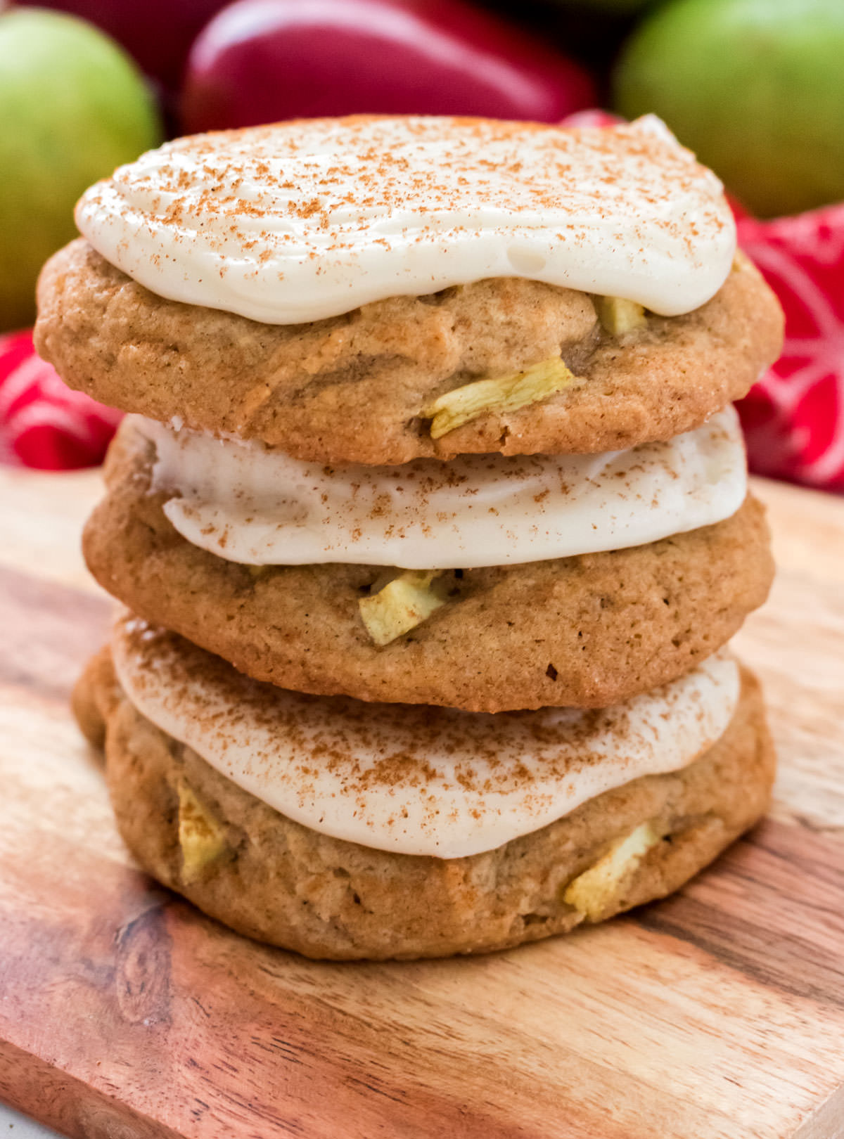 Closeup on a stack of three Apple Cookies with Cream Cheese Frosting sitting on a cutting board in front of a red towel and red and green apples.