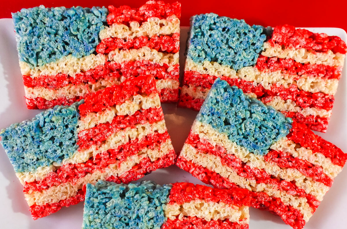 Closeup on a plate of Red White and Blue 4th of July Rice Krispie Treats sitting on a white serving platter that is sitting on a red table.