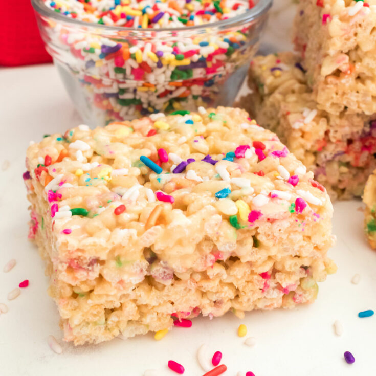 Square image on a single Cake Batter Rice Krispie Treats sitting on a white table in front of a small glass bowl filled with sprinkles.