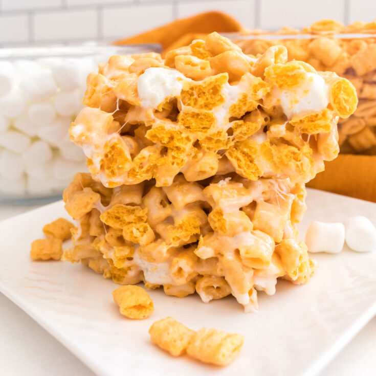 Square image of two Cap'n Crunch Marshmallow Treats sitting on a white plate in front of a clear bowl filled with Mini Marshmallows.