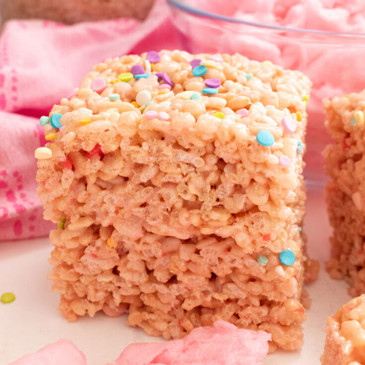 Square image of two Cotton Candy Rice Krispie Treats sitting on a white table surrounded by sprinkles, cotton candy and a pink kitchen towel.