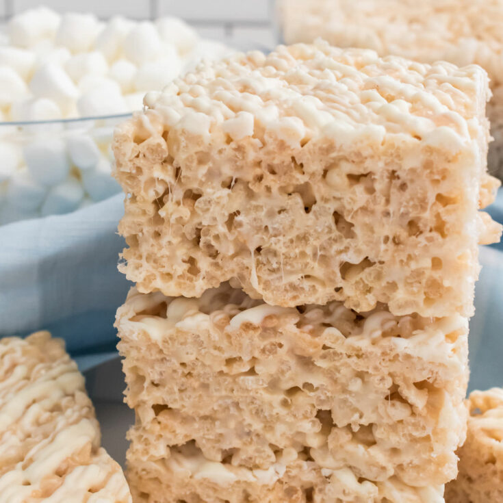 Square image of a stack of two White Chocolate Rice Krispie Treats sitting on a white table in front of a bowl of Mini Marshmallows and a blue kitchen towel.