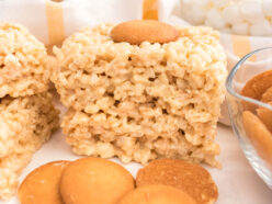 A stack of two Banana Pudding Rice Krispie Treats sitting on a white table surrounded by Nilla Wafers and bowls of cereal and marshmallows.