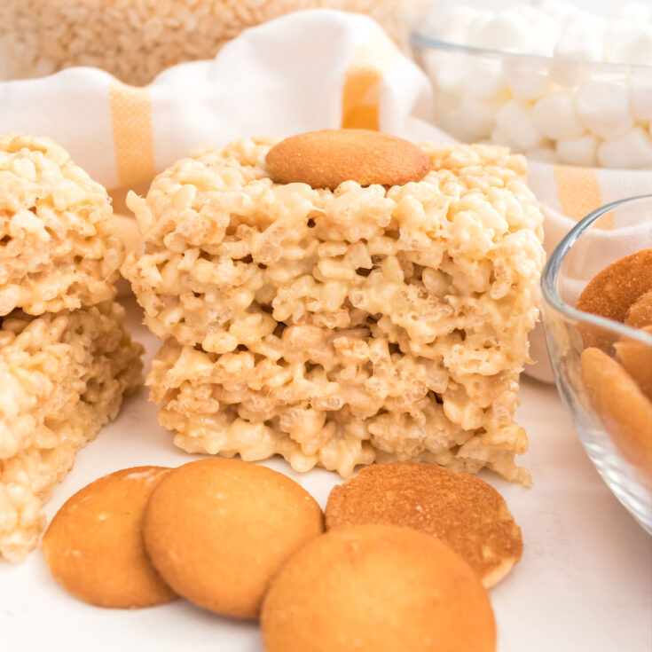 A stack of two Banana Pudding Rice Krispie Treats sitting on a white table surrounded by Nilla Wafers and bowls of cereal and marshmallows.