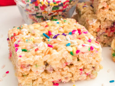 A single Cake Batter Rice Krispie Treat sitting on a white table surrounded by sprinkles.