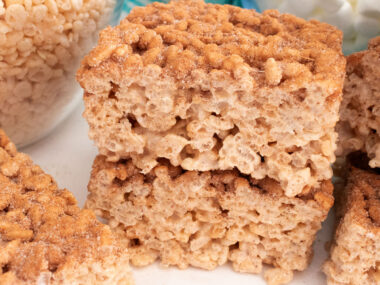 A stack of two Churro Rice Krispie Treats sitting on a white table in front of glass bowls filled with marshmallows and cereal.