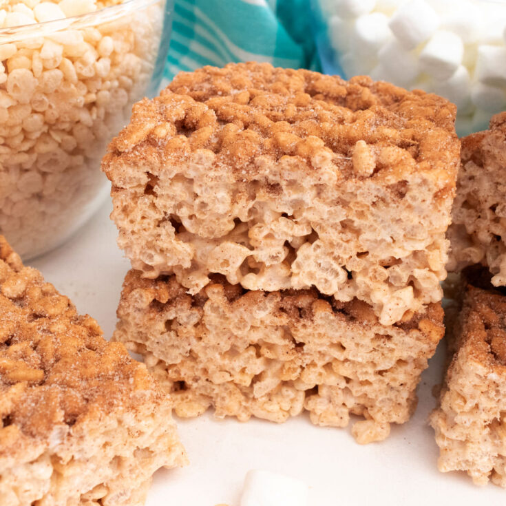 A stack of two Churro Rice Krispie Treats sitting on a white table in front of glass bowls filled with marshmallows and cereal.