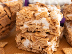 Two Cinnamon Toast Crunch Marshmallow Treats sitting on a cutting board surrounded by cereal and marshmallows.