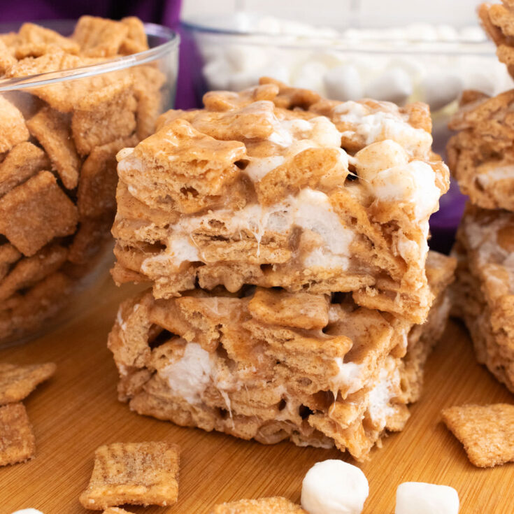 Two Cinnamon Toast Crunch Marshmallow Treats sitting on a cutting board surrounded by cereal and marshmallows.