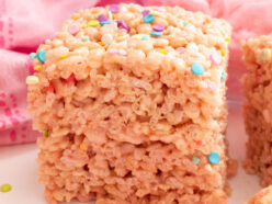 Square image of a stack of two Cotton Candy Rice Krispie Treats sitting on a white table surrounded by Pink Cotton Candy.