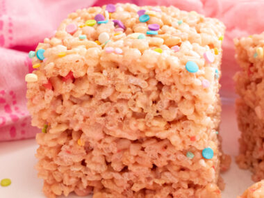 Square image of a stack of two Cotton Candy Rice Krispie Treats sitting on a white table surrounded by Pink Cotton Candy.