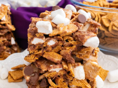 Two Golden Grahams S'mores Marshmallow Treats sitting on a white dessert plate surrounded by glass bowls filled with marshmallows and cereal.