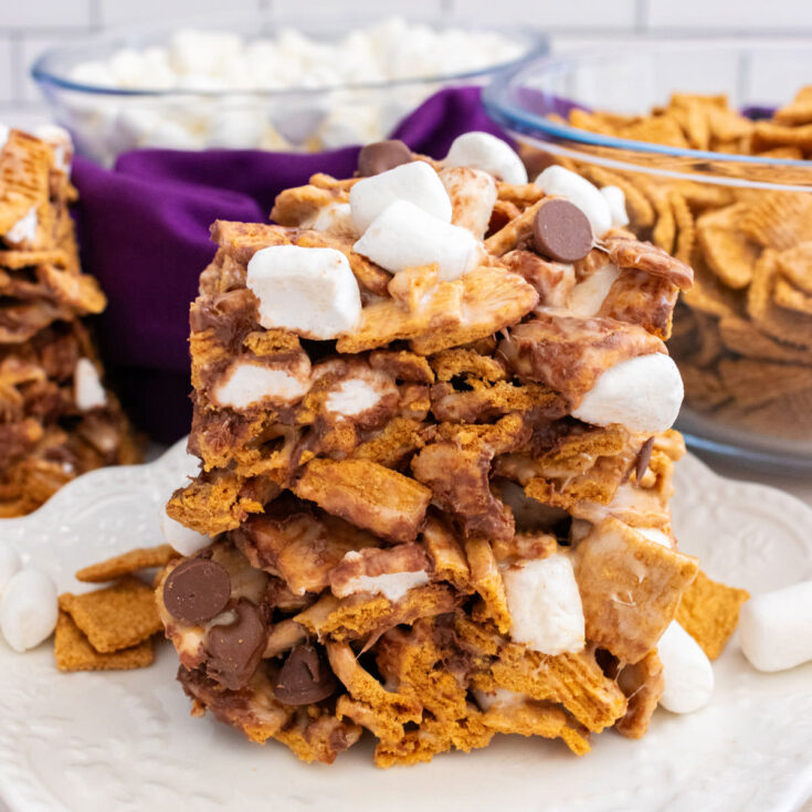 Two Golden Grahams S'mores Marshmallow Treats sitting on a white dessert plate surrounded by glass bowls filled with marshmallows and cereal.