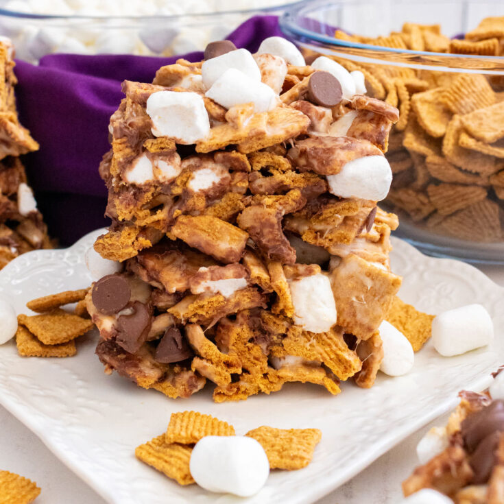 Two Golden Grahams S'mores Marshmallow Treats sitting on a white dessert plate surrounded by glass bowls filled with marshmallows and cereal.