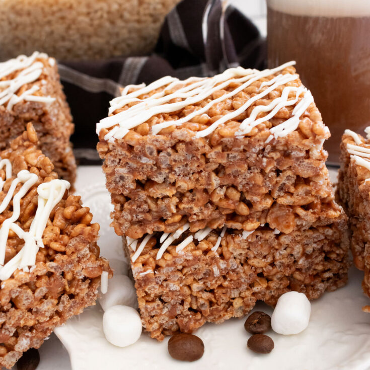 A stack of two Mocha Rice Krispie Treats sitting on a white dessert plate next to a glass cup filled with Mocha Coffee.