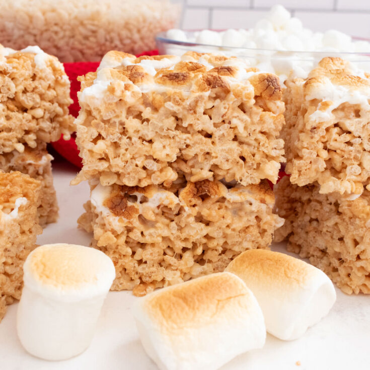 A batch of Toasted Marshmallow Rice Krispie Treats sitting on a white table surrounded by roasted marshmallows.