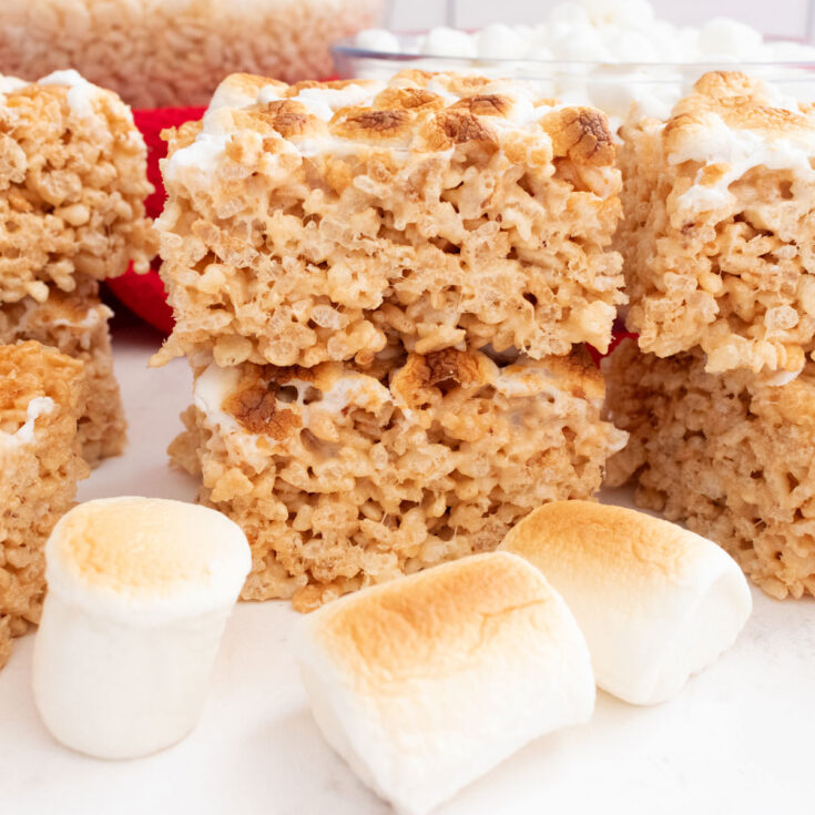 Stacks of Toasted Marshmallow Rice Krispie Treats sitting on a white table in front of three Roasted Marshmallows.