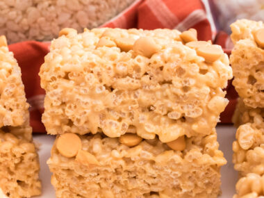 A stack of two Butterscotch Rice Krispie Treats sitting on a white table surrounded by Butterscotch Chips and Mini Marshmallows.