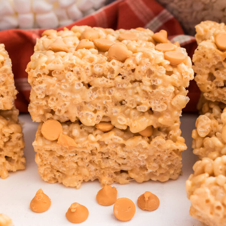 A stack of two Butterscotch Rice Krispie Treats sitting on a white table surrounded by Butterscotch Chips and Mini Marshmallows.