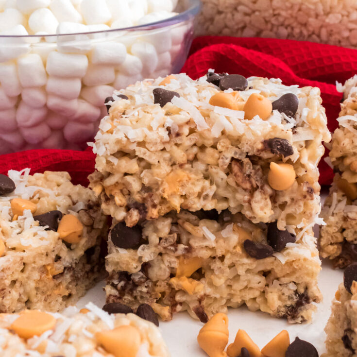 A stack of two Magic Bar Rice Krispie Treats sitting on a white table surrounded by Butterscotch Chips, Chocolate Chips and Coconut.