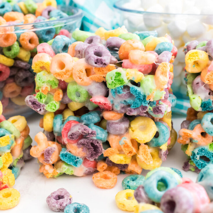 Close-up on a stack of two Froot Loops Marshmallow Treats sitting on a white table in front of glass bowls filled with Froot Loop Cereal and Mini Marshmallows.