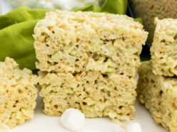 Closeup on a stack of two Pistachio Rice Krispie Treats sitting on a white table in front of bowls of Mini Marshmallows and Rice Krispies Cereal.