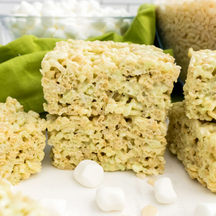 Closeup on a stack of two Pistachio Rice Krispie Treats sitting on a white table in front of bowls of Mini Marshmallows and Rice Krispies Cereal.