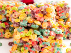 Closeup on a stack of two Trix Marshmallow Treats sitting on a white table in front of glass bowls filled with Trix Cereal and Mini Marshmallows.