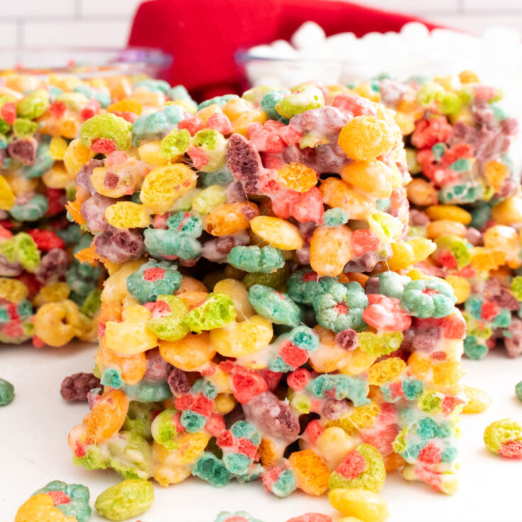 Closeup on a stack of two Trix Marshmallow Treats sitting on a white table in front of glass bowls filled with Trix Cereal and Mini Marshmallows.